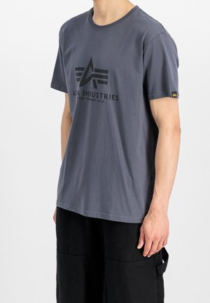 Mężczyzna noszący szarą koszulkę z krótkim rękawem z czarnym logo i napisem "Alpha Industries", w połączeniu z czarnymi spodniami typu cargo.
