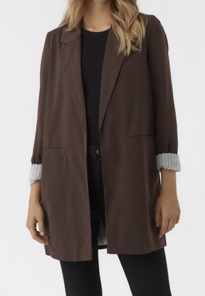 Femme portant un blazer marron ouvert avec des poignets rayés retroussés sur un haut noir et un pantalon noir, montrée du cou jusqu'à mi-cuisse.