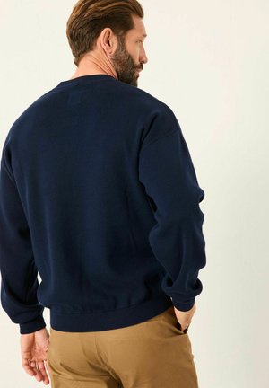 Man with beard and short hair wearing a navy sweater and tan pants facing away against a plain light background.