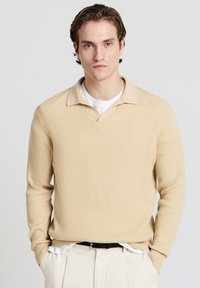 Beige Rippstrickpullover mit V-Ausschnitt und Kragen. Lange Ärmel, lockere Passform, kombiniert mit einem weißen Hemd, mit strukturierter Oberfläche.
