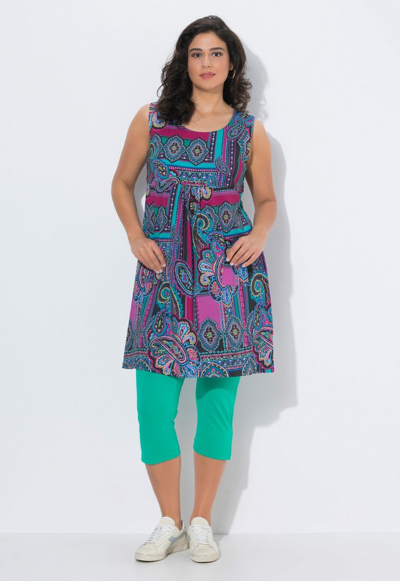 Femme debout portant une tunique sans manches à motifs colorés avec des poches à l'avant, des leggings verts raccourcis et des baskets blanches sur un fond uni.