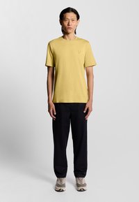 Gul bomulds T-shirt med korte ærmer og rund hals, der har et lille logo. Parret med sorte løstsiddende bukser og beige sneakers.