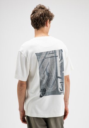 Man met krullend haar draagt wit shirt met korte mouwen met grote abstracte grijs-zwarte afbeelding op de rug, staand tegen een effen achtergrond.