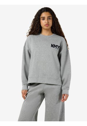Femme aux longs cheveux bouclés portant un sweat-shirt gris ample avec des lettres "NMY" et un pantalon gris large assorti.