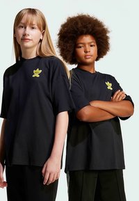 Juodi trumpomis rankovėmis marškinėliai su geltonos spalvos lapo dizainu ant krūtinės. Modeliai stovi šalia vienas kito, turėdami skirtingus šukuosenų ir apačių variantus.