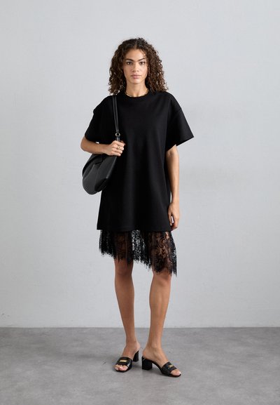 Fekete oversized pólóruha csipkés szegéllyel, laza szabású. Fekete, blokk-sarkú szandálokkal és fekete válltáskával párosítva.