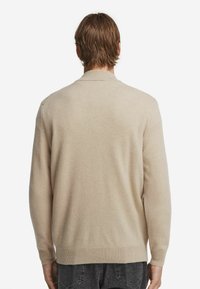 Mann mit kurzen braunen Haaren, der einen beigen Langarmpullover und dunkelgraue Jeans trägt, von hinten vor einem weißen Hintergrund gezeigt.