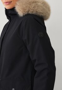Giacca parka nera con cappuccio in pelliccia staccabile di colore marrone chiaro, chiusura frontale con zip, grandi tasche laterali e patch logo sulla manica sinistra.