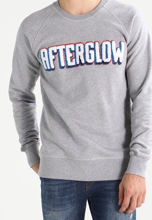 Mann trägt ein graues Sweatshirt mit strukturiertem "AFTERGLOW"-Text in Weiß, Blau und Rot über der Brust, kombiniert mit blauen Jeans.