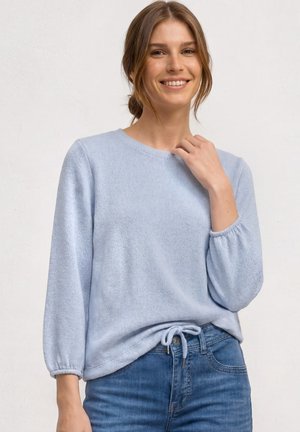 Junge Frau lächelt, trägt einen hellblauen Langarmpullover mit Kordelzug am Saum und blaue Jeans, steht vor einem einfarbigen hellen Hintergrund.