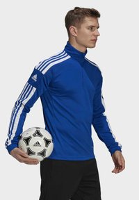 Blauwe sportpullover met een ritskraag, voorzien van witte strepen op de mouwen en een Adidas-logo. Het model houdt een zwart-wit voetballetje vast.