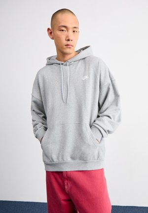 Sudadera gris con bolsillo frontal y capucha con cordón. Presenta un pequeño logo blanco de Nike en el pecho. La tela parece suave y acogedora.