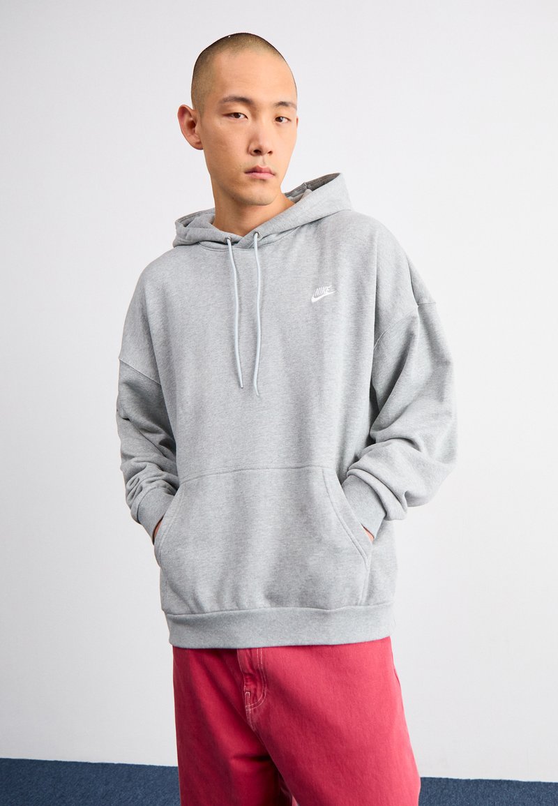 Szary hoodie z kieszenią z przodu i kapturem ze sznurkiem. Na klatce piersiowej znajduje się małe białe logo Nike. Materiał wydaje się miękki i przytulny.