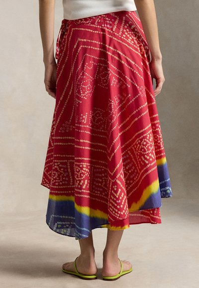 Nainen pukeutuneena punaiseen, kuvioituun midihameeseen, jossa on keltainen ja sininen reunus, yhdistettynä ohuilla remmeillä varustettuihin vihreisiin sandaaleihin, kuvattuna takaa.