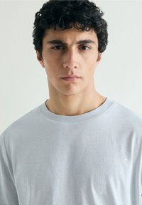 Joven con cabello oscuro y rizado lleva una sudadera gris claro con el texto "Creative Studio" y un logo de calavera, mirando hacia adelante con expresión neutral.