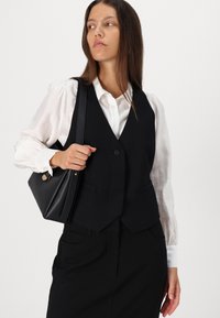 Black tailored vest with two front pockets, worn over a white blouse. The model holds a black leather handbag with a gold accent.