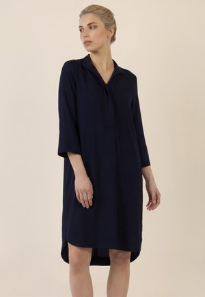 Frau trägt ein locker sitzendes, knielanges navyblaues Kleid mit dreiviertel Ärmeln, steht vor einem schlichten beigen Hintergrund und blickt nach links.