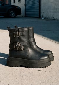 Paire de bottines noires en cuir avec semelles épaisses crantées et brides à double boucle sur un trottoir en béton en plein jour.