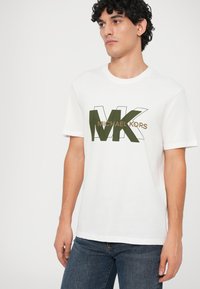 Vīrietis baltā T-kreklā ar īsām piedurknēm, uz kura ir zaļš un brūns "MK Michael Kors" logo, ģērbies zilos džinsos, skatās uz sāniem uz balta fona.