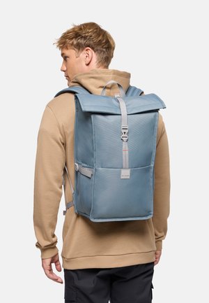 Jonge man met een beige hoodie en zwarte broek, met een grote blauwe rechthoekige rugzak die is bevestigd met een centrale gesp.