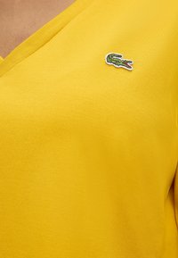 T-shirt jaune à col en V avec un petit logo crocodile vert brodé sur la poitrine côté gauche.