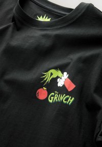 Czarna koszulka z zieloną ręką trzymającą czerwony ornament, z napisem "The Grinch" w zielonym kolorze poniżej. Wykonana z miękkiej bawełny.