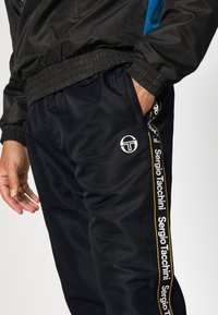 Fekete sportnadrág, texturált felülettel, fehér logóval és oldalsávokkal, melyeken sárga és fehér "Sergio Tacchini" felirat található.