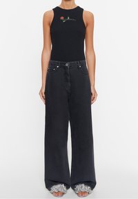 Nainen, joka on pukeutunut mustaan, hihattomaan, resorilliseen toppiin, jossa on pieni kukkakuvio, leveälahkeisiin mustiin farkkuihin ja hopeisiin kukkakuvioituihin sandaaleihin.