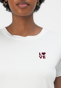 White cotton t-shirt featuring a burgundy embroidered "LOVE" logo with a heart on the left chest area. Short sleeves with a rounded neckline.
