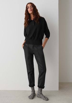 Black pullover with a collar and button detail, paired with fitted black trousers featuring side seams. Gray patterned socks are visible.