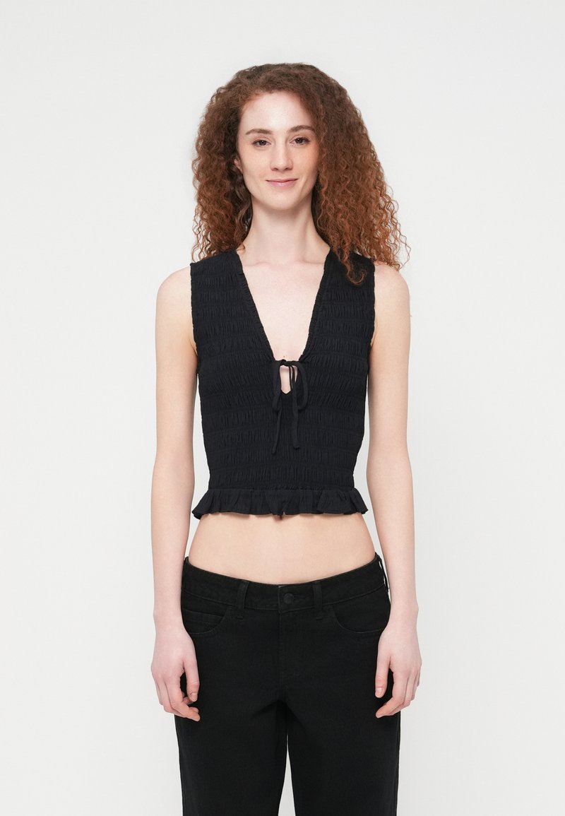 Μαύρο κοντό τοπ από υφασμάτινη υφή με βαθύ V-λαιμόκοψη, λεπτομέρεια δέσιμου μπροστά και βολάν στο τελείωμα. Συνδυάζεται με μαύρο παντελόνι ψηλόμεσο.