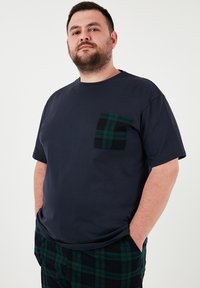 Granatowa bawełniana koszulka z kieszonką w zielono-czarną kratkę. Prosty kroj z okrągłym dekoltem i krótkimi rękawami. Luźny fason.