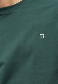 Zielona bawełniana koszulka z okrągłym dekoltem, z małym białym haftowanym napisem "II" na piersi. Miękka w dotyku i gładka w wykończeniu.