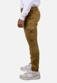 Pantalones cargo color bronce con puños ajustados, bolsillos laterales y traseros, y una etiqueta. Combinados con zapatillas deportivas blancas.