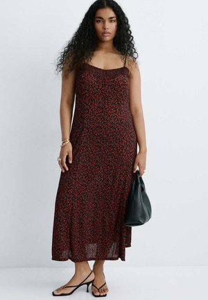 Femme aux longs cheveux bouclés portant une robe midi sans manches noire et rouge à fleurs, des sandales noires à talons et tenant un sac à main noir.