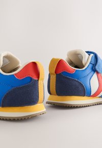 Paire de baskets colorées pour tout-petits avec des panneaux bleus, rouges, jaunes et blancs, dotées de sangles à scratch et de semelles en caoutchouc texturées.
