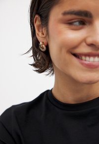 Guldtonade örhängen med en texturerad virveldesign med kristalldekorationer. Modellen bär en svart t-shirt som framhäver örhängena på ett framträdande sätt.