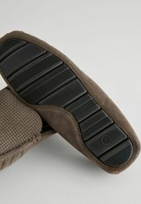 Brune ruskinds-slipper med en struktureret overflade og en sort gummisål med horisontale riller for bedre greb. Syede detaljer langs kanten.