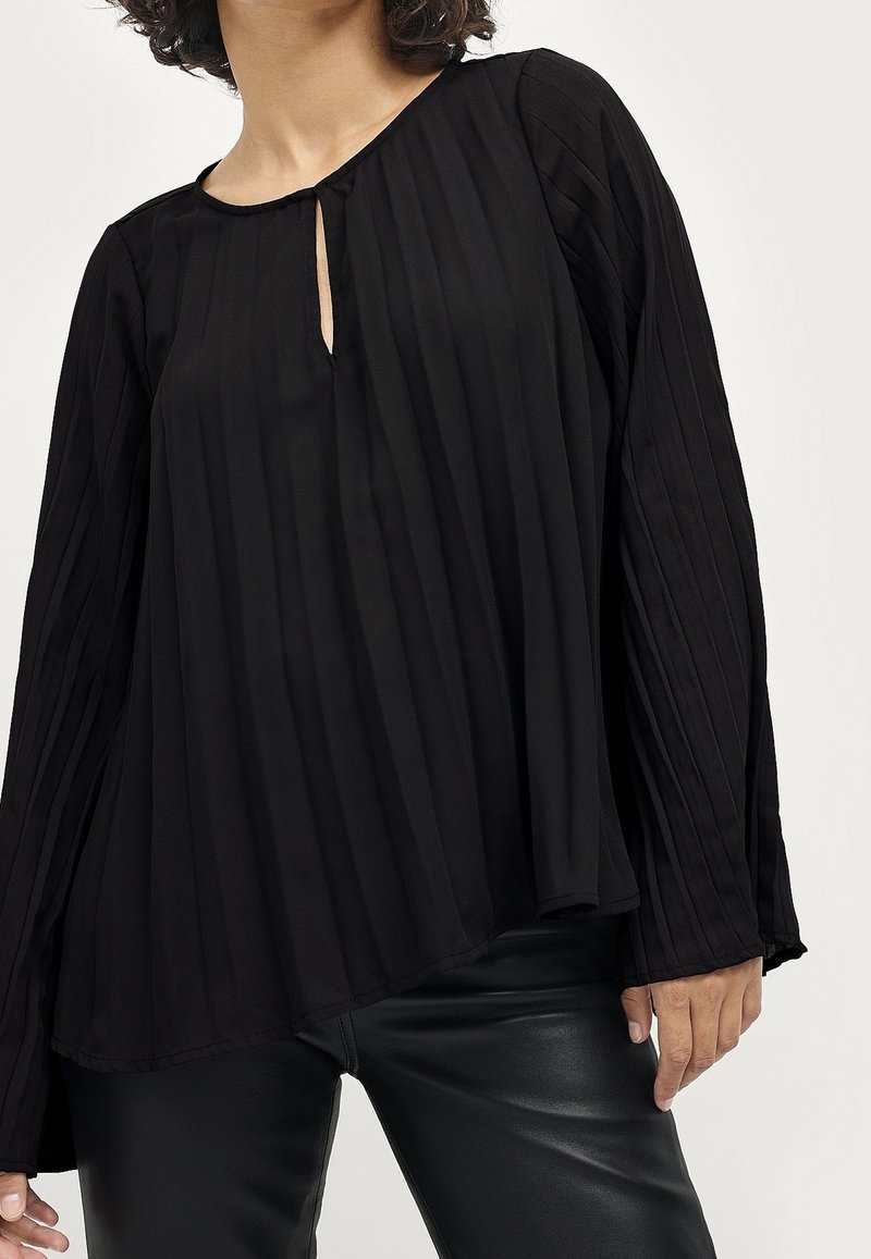 Femme portant un chemisier plissé noir à manches longues avec un petit décolleté en avant et un pantalon en cuir noir.