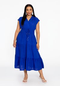 Blaues, gestuftes Midi-Kleid mit einem Wickel-Oberteil, kurzen Kapp�ärmel, Kordelzug-Taillierung und strukturiertem Stoff. Getragen mit weißen Sandalen.