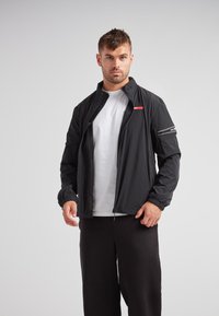 Sort letvægtsjakke med lynlås foran, elastiske manchetter og en rød accentstribe på brystet. Stylingset med en hvid t-shirt underneath.