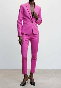 Traje fucsia con una chaqueta ajustada y pantalones de corte slim. La textura es suave, con un cierre de un solo botón. Combinado con tacones negros.