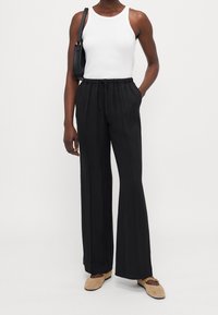 Mustat, leveälahkeiset housut, joissa on naru vyötäröllä ja pehmeä tekstuuri, yhdistetty valkoiseen hihattomaan toppiin sekä beigeihin kenkiin. Yksinkertainen muotoilu.