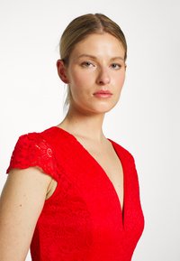 Vestido vermelho de renda com um decote em V profundo e mangas curtas com borda recortada. O padrão floral é intricado, criando uma aparência texturizada.