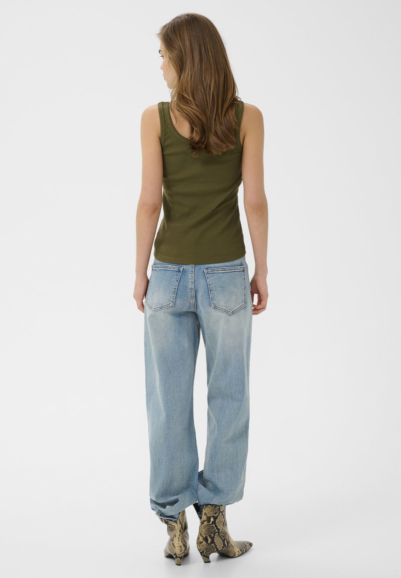 Jeune femme aux longs cheveux portant un débardeur vert olive, un jean bleu clair ample et des bottines à motif peau de serpent, debout de dos.