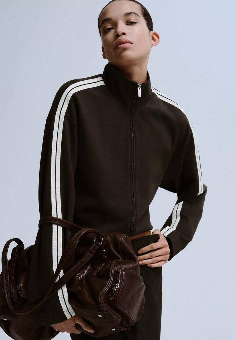 Brun lynlåsjakke med hvide striber på ærmerne; glat stof. Modellen holder en mørkebrun håndtaske med lynlåse og afrundet form.