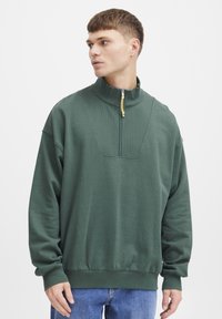 Sudadera verde con cremallera y cuello alto, panel frontal texturizado, puños y bajo de canalé, que presenta un detalle de logo circular en el pecho.
