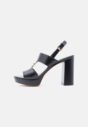 Sandalo con tacco alto in pelle nera con suola platform, cinturini larghi, dettaglio fibbia dorata e cinturino regolabile alla caviglia su sfondo bianco.