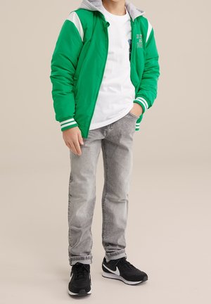 Zielona pikowana kurtka bomberka z białymi akcentami i szarym kapturem, noszona na białej koszulce, w połączeniu z szarymi jeansami i czarnymi trampkami.