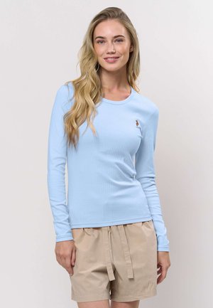 Frau mit langen blonden Haaren, die ein hellblaues Langarmshirt und beige Shorts trägt und vor einem schlichten hellen Hintergrund steht.